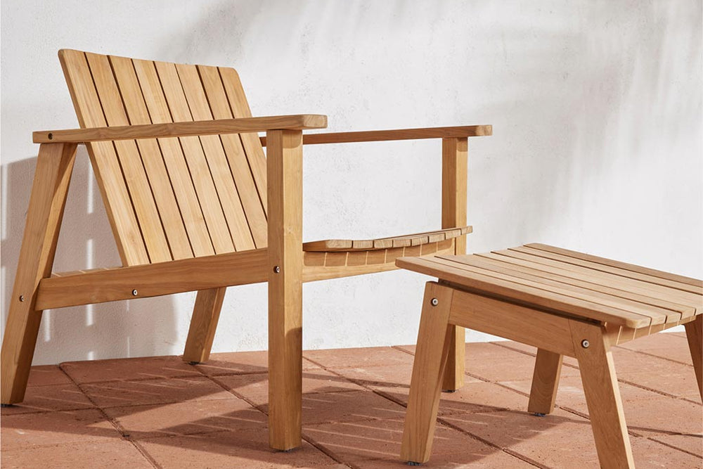 Neighbor Low Chair in natural teak slatted frame with matching Low Ottoman on a terracotta tile patio against a white stucco wall