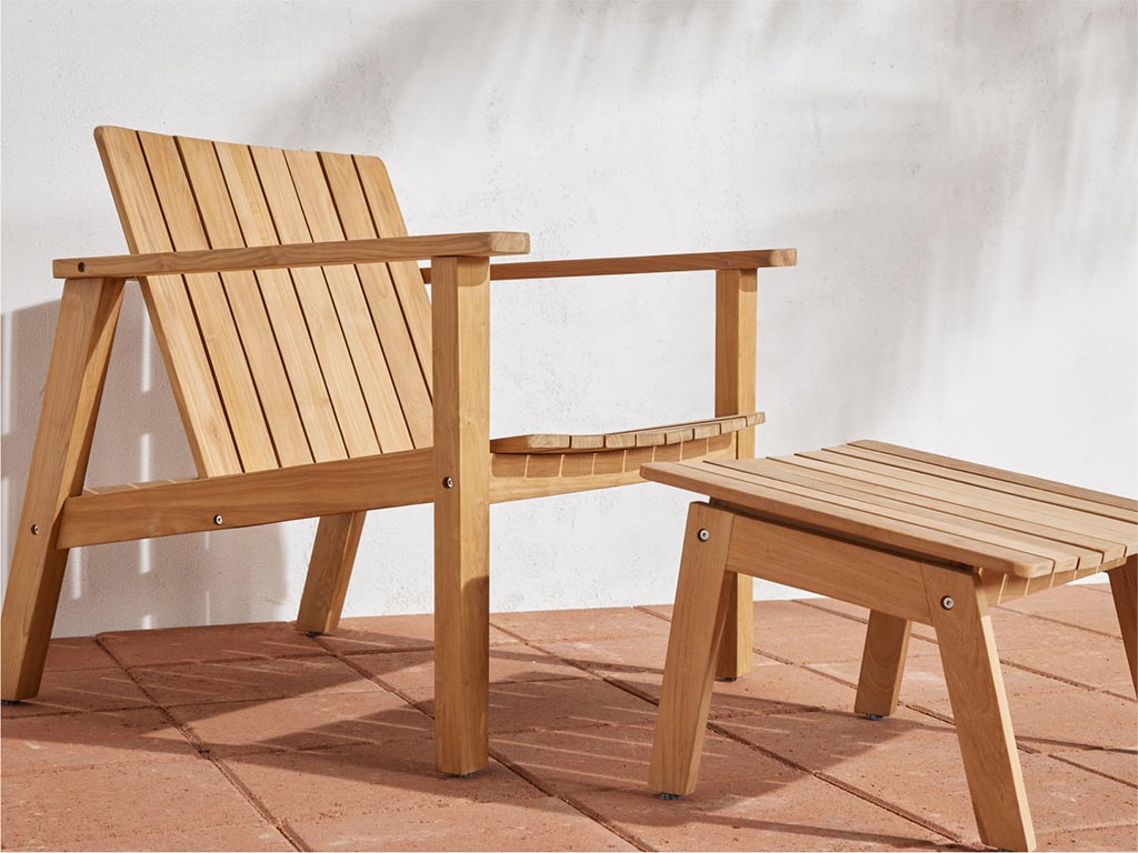 Neighbor Low Chair in natural teak slatted frame with matching Low Ottoman on a terracotta tile patio against a white stucco wall