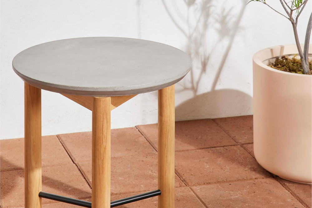 Neighbor Round Side Table with concrete top and teak tripod legs on terracotta tile, large pink planter with bare tree beside it