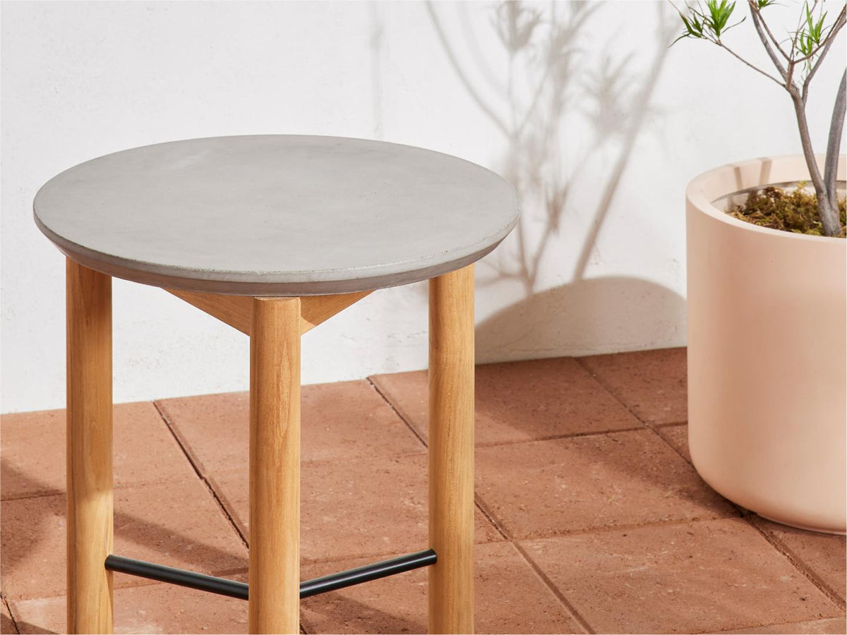 Neighbor Round Side Table with concrete top and teak tripod legs on terracotta tile, large pink planter with bare tree beside it