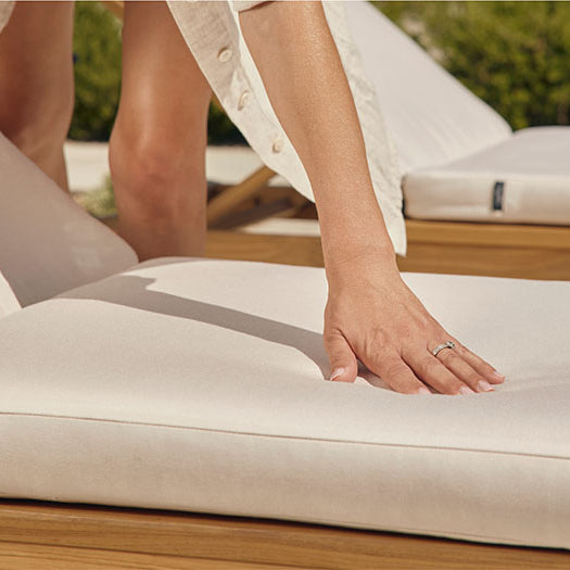 Person pressing a hand into a thick white Neighbor outdoor cushion with Neighbor label tag visible, teak chaise lounges visible in background