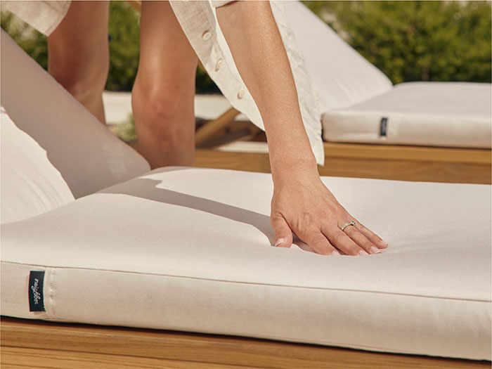 Person pressing a hand into a thick white Neighbor outdoor cushion with Neighbor label tag visible, teak chaise lounges visible in background