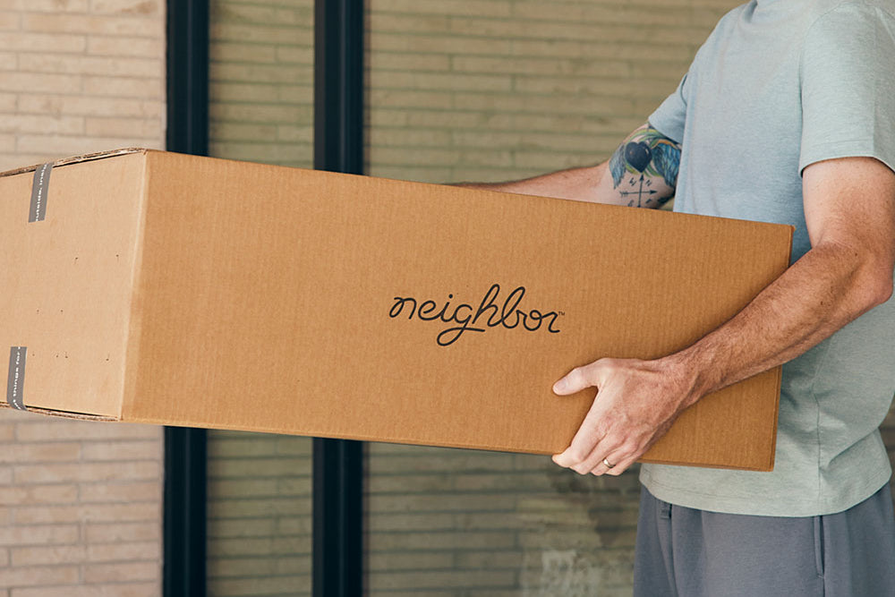 Man carrying a Neighbor-branded cardboard shipping box near a brick building