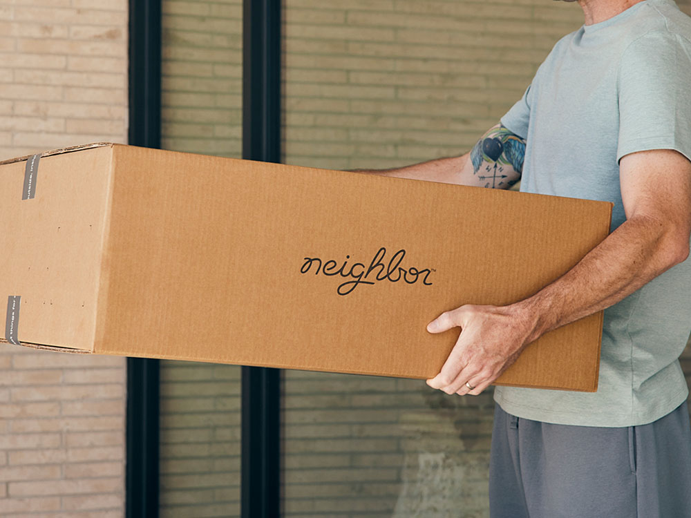 Man carrying a Neighbor-branded cardboard shipping box near a brick building