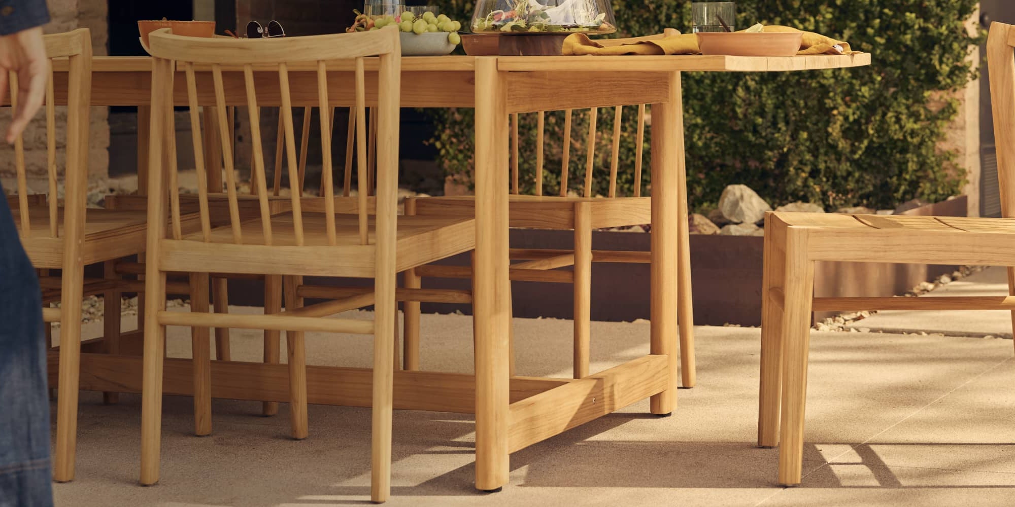 Windsor-styled teak wood dining chair next to a teak dining table on a warm outdoor patio with someone cropped off left edge, walking into frame.