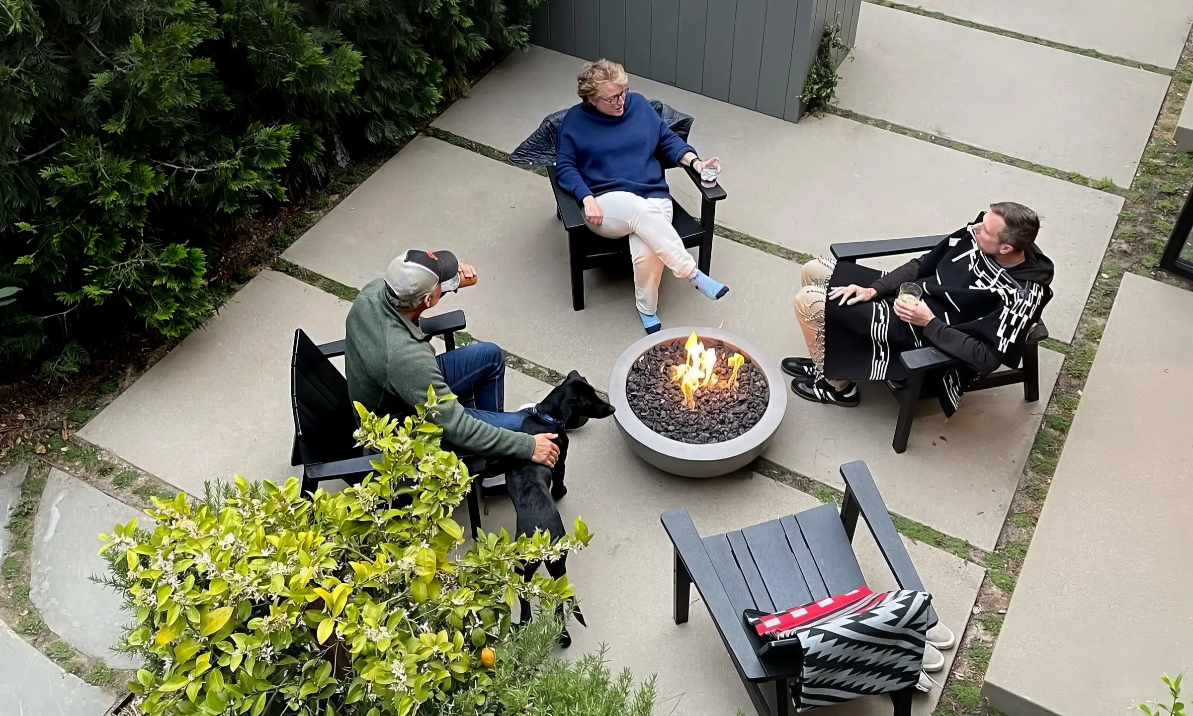 Three people seated on charcoal outdoor chairs around fire table on concrete patio in lush garden setting