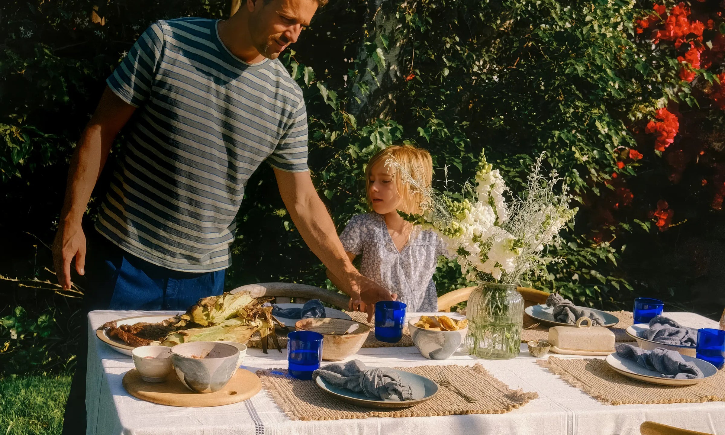 Man and child at outdoor dining table with wooden furniture, food, and fresh flowers in lush garden
