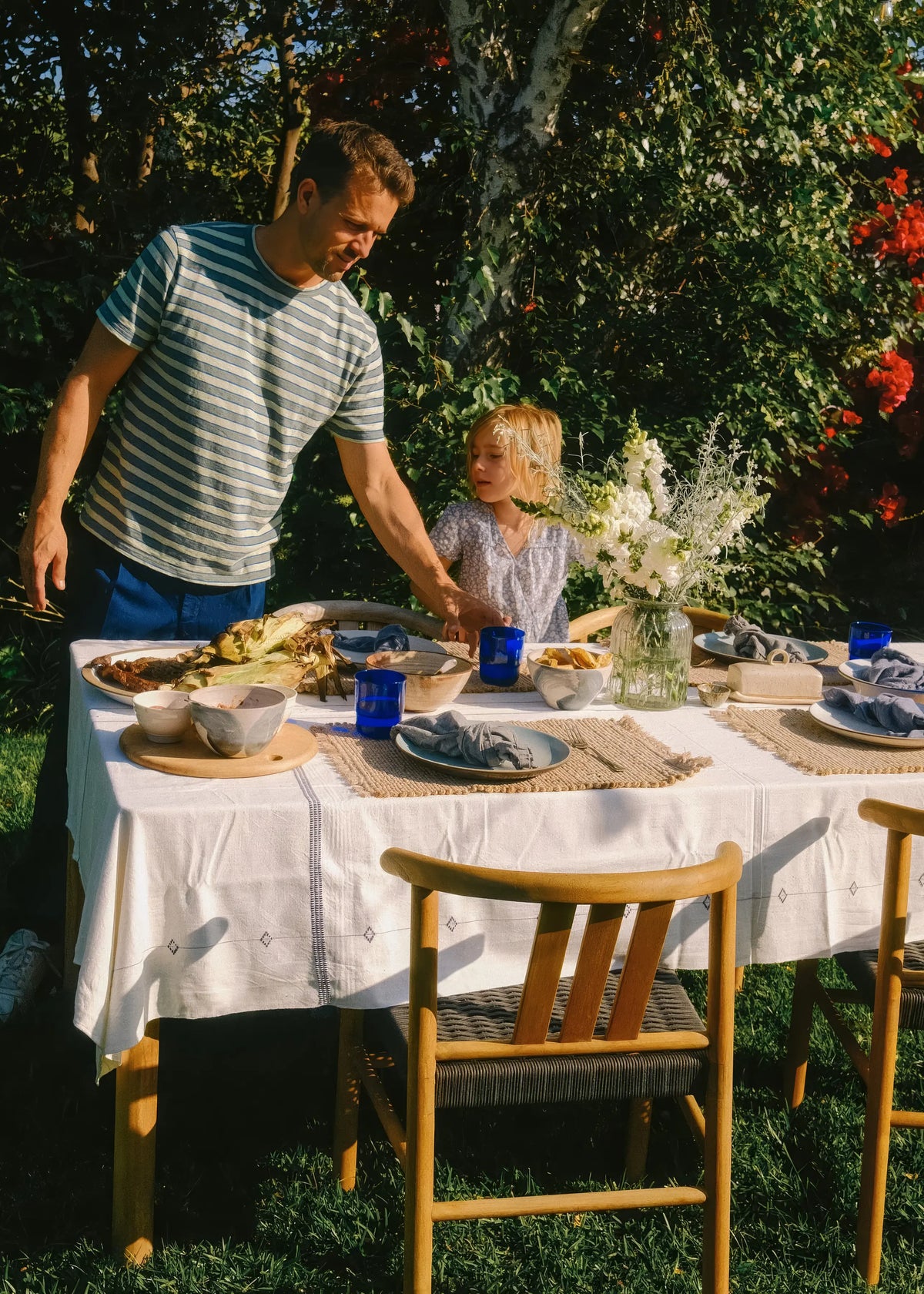 Man and child at outdoor dining table with wooden furniture, food, and flowers in lush garden setting