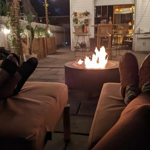 Customer photo - two people's feet propped up near a Neighbor round fire table at night, backyard patio with string lights