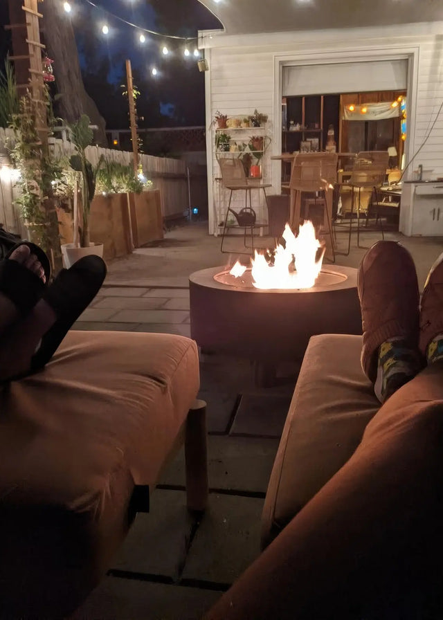 Customer photo - two people's feet propped up near a Neighbor round fire table at night, backyard patio with string lights