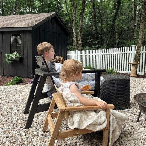 Customer photo - two young children sitting in Neighbor Low Chairs in natural teak on a gravel backyard patio, dark garden shed and white fence behind
