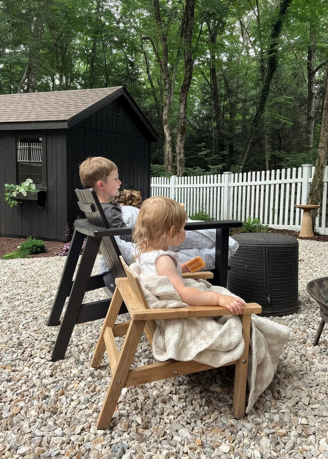 Customer photo - two young children sitting in Neighbor Low Chairs in natural teak on a gravel backyard patio, dark garden shed and white fence behind